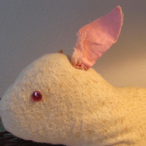 May include: A handmade, off-white bunny rabbit with a pink inner ear and a red bead eye. The bunny is made of a textured fabric and is a charming, rustic decoration.