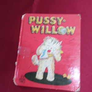 May include: A vintage children's book titled "Pussy Willow" with a red cover. The cover features a cartoon illustration of a white cat-like character holding a yellow object and wearing a blue bib. The title is in yellow letters.
