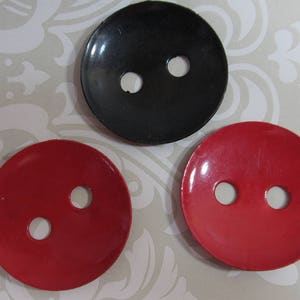 May include: Three round buttons: one black and two red. Each button has two holes for sewing. The buttons are on a patterned, light-colored surface. These buttons are suitable for various sewing projects.