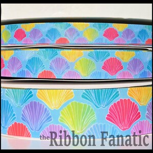 May include: A spool of light blue grosgrain ribbon featuring a repeating pattern of colorful seashells in shades of pink, purple, yellow, and teal. The text the Ribbon Fanatic is printed in dark gray at the bottom of the ribbon spool.