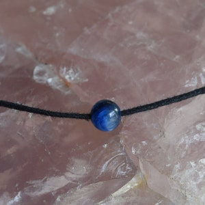 Peut inclure: Un délicat collier en cordon noir avec une seule perle bleue ronde. Le collier est présenté sur un grand cristal rose à texture rugueuse. Un petit pot de plante flou est visible en arrière-plan.