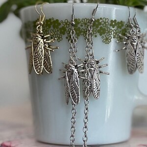 Könnte beinhalten: Eine weiße Keramiktasse mit grünen floralen Details enthält grüne Pflanzen und mehrere Paare von Ohrringen in Insektenform. Die Ohrringe sind silber- und goldfarben mit aufwendigen Details. Ein schwarzes Ohrring-Display-Rack befindet sich im Hintergrund.
