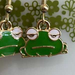 May include: A pair of gold dangle earrings with green frog charms. The frogs have white eyes and a green body with a gold outline.