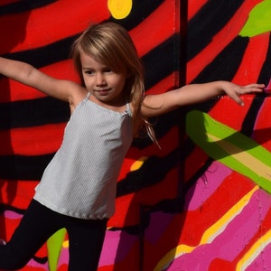 May include: A young girl wearing a striped tank top and black leggings poses in front of a colorful wall with pink, red, yellow, and green stripes. She is wearing pink rain boots.