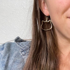 May include: Gold cat-shaped earrings with a simple design. The earrings are hanging from the ears of a person with long brown hair with green highlights.