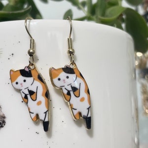 May include: A pair of gold dangle earrings featuring a cartoon calico cat design. The cat is orange, black, and white with a pink nose and white whiskers.