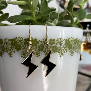May include: A pair of gold earrings with black lightning bolt charms. The earrings are hanging from a white ceramic mug with a green floral pattern.