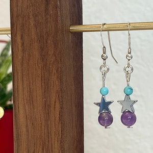 May include: Three pairs of dangle earrings with silver hooks. Each earring features a star charm, a turquoise bead, a yellow bead, and a purple bead.