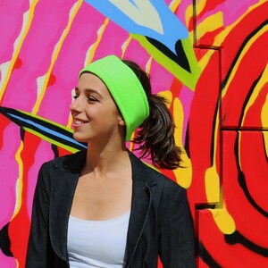 May include: A woman with long brown hair wearing a bright green headband and a black blazer. She is standing in front of a colorful graffiti wall.