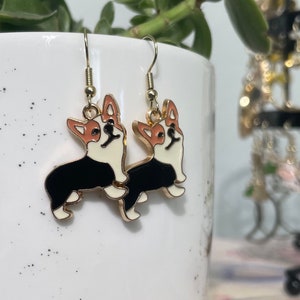May include: Gold dangle earrings featuring a black and white corgi dog design. The corgis are facing forward with their tongues sticking out.