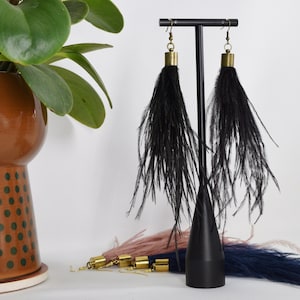 May include: A pair of long black feather earrings with gold accents hanging from a black stand. The earrings are approximately 6 inches long.
