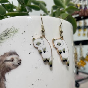 Può includere: Un paio di orecchini pendenti dorati con ciondoli a forma di gatto bianco. I gatti sono rivolti in avanti con le zampe alzate e le code arrotolate attorno ai loro corpi.