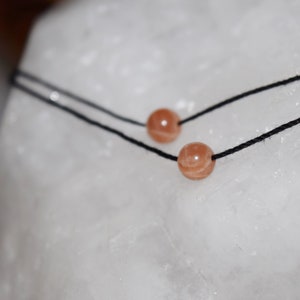 Peut inclure: Gros plan sur un collier délicat composé de deux cordons noirs. Chaque cordon est orné d'une petite pierre précieuse ronde de couleur pêche. Le collier est présenté sur un fond blanc texturé, soulignant sa simplicité.