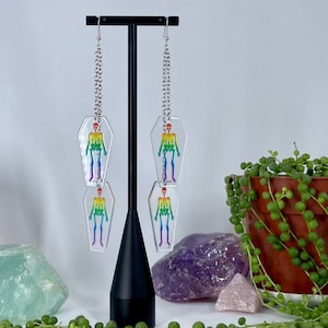 May include: Rainbow skeleton earrings in a coffin shape, hanging from silver chains. The earrings are displayed on a black stand with crystals and greenery.
