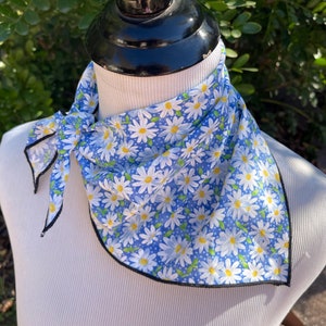 May include: A blue bandana with a white daisy pattern and a black border is tied around a mannequin's neck. The bandana is made of a lightweight fabric and is knotted at the front. The background is a blurred view of green foliage.