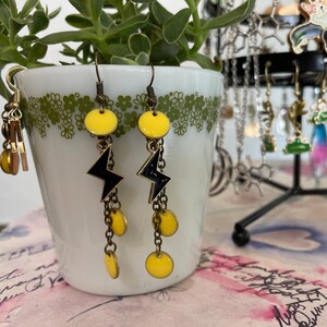 May include: A pair of dangle earrings with black lightning bolt charms and yellow round beads. The earrings are hanging from a white ceramic planter with green floral designs.