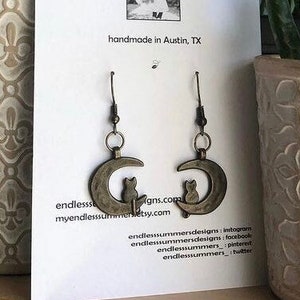 Può includere: Un paio di orecchini dorati con un design a forma di luna crescente e una piccola silhouette di gatto su ogni orecchino. Gli orecchini sono fatti a mano ad Austin, Texas. La carta include anche il nome del marchio "endless.summers" e le informazioni di contatto del negozio.