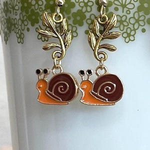 May include: Gold-colored snail earrings with orange and brown enamel details. The earrings have a leaf design above the snail charm and are displayed on a white mug with a green floral pattern. The earrings are a whimsical accessory.