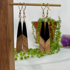 Może przedstawiać: Para długich, drewnianych kolczyków z czarnymi końcówkami z żywicy. Kolczyki wiszą na złotych haczykach.
