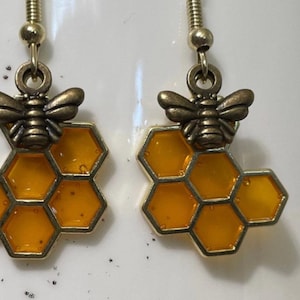 May include: A pair of gold-toned earrings featuring a honeycomb design with a small bee charm on top. The honeycomb is a bright yellow color.