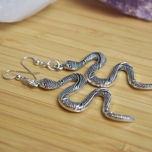 May include: Silver-toned snake earrings with detailed scales and curved bodies. The earrings are displayed on a wooden surface, showcasing their intricate design and metallic sheen. The snake earrings are a unique jewelry item.