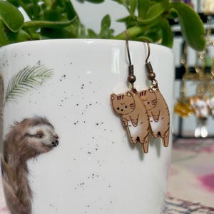 Puede incluir: Un par de pendientes colgantes con un diseño de gato marrón con vientre blanco. El gato está colgando boca abajo con las patas en el aire. Los pendientes están hechos de metal y tienen un acabado dorado.