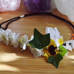 May include: A black headband with white flowers and a yellow sunflower, accented with green leaves.