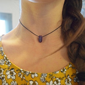 Peut inclure: Un collier ras du cou minimaliste avec un petit pendentif en pierre précieuse violette polie. Le pendentif est suspendu à un fin cordon noir. Le modèle porte un haut jaune à imprimé floral.