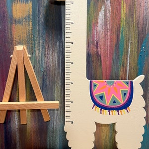 May include: A wooden llama-shaped growth chart with a ruler printed on the neck. The llama is white with a colorful blanket design and a pink and blue headband. The chart measures in inches. A small wooden easel is to the left.