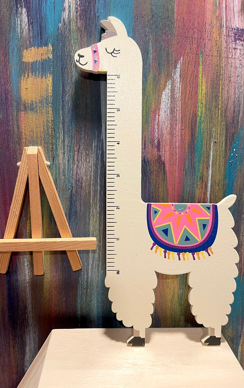 May include: A wooden llama-shaped growth chart with a ruler printed on the neck. The llama is white with a colorful blanket design and a pink and blue headband. The chart measures in inches. A small wooden easel is to the left.
