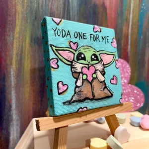 May include: A small, square canvas painting on a wooden easel. The painting features a cartoon depiction of Grogu, holding a pink heart, with the text "YODA ONE FOR ME" on a light blue background. Pink hearts are scattered around the character.