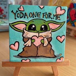 May include: A small, square canvas painting on a wooden easel. The painting features a green character with large eyes and the text "YODA ONE FOR ME". Pink hearts are scattered around the character.