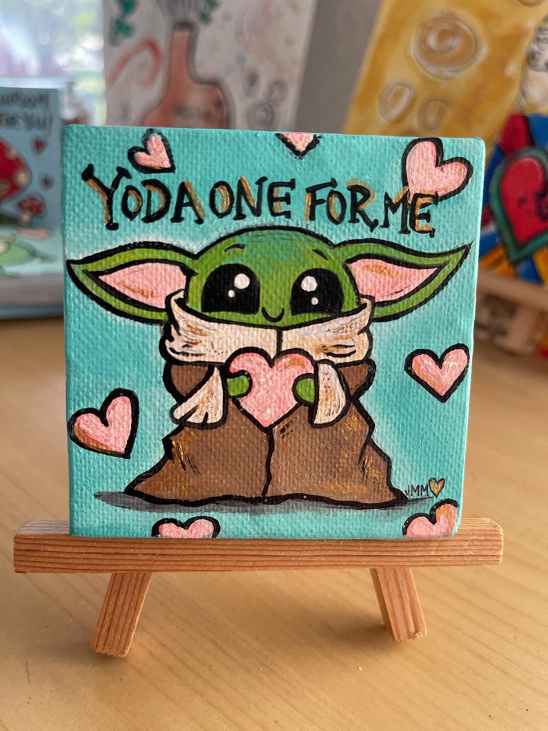May include: A small, square canvas painting on a wooden easel. The painting features a green character with large eyes and the text "YODA ONE FOR ME". Pink hearts are scattered around the character.