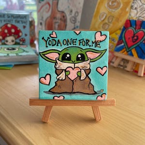 May include: A small square canvas painting on a wooden easel. The painting features a green character holding a pink heart, with the text "YODA ONE FOR ME" above. Pink hearts are scattered around the character against a teal background.
