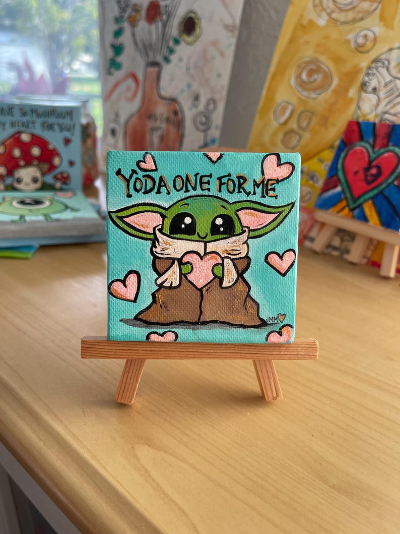 May include: A small square canvas painting on a wooden easel. The painting features a green character holding a pink heart, with the text "YODA ONE FOR ME" above. Pink hearts are scattered around the character against a teal background.