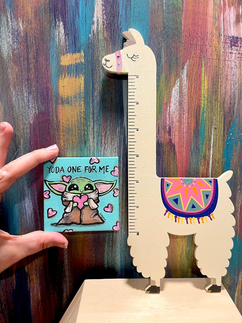 May include: A small square canvas painting featuring a cartoon character holding a heart, with the text "YODA ONE FOR ME". A wooden llama-shaped ruler stands next to the painting. The ruler has a colorful design on its back and measures up to 6 inches.