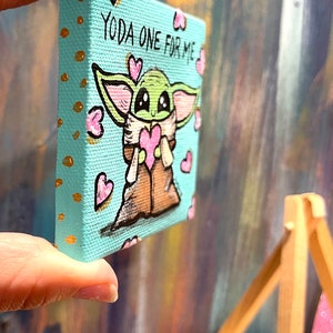 May include: A small, square canvas art piece featuring a cartoon character resembling Yoda, holding a pink heart. The background is a light blue with gold dots and pink hearts. Text reads "YODA ONE FOR ME."