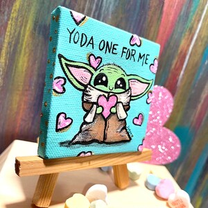 May include: A small, square canvas painting on a wooden easel. The artwork features a green character holding a pink heart, surrounded by pink hearts, with the text "YODA ONE FOR ME" on a light blue background.