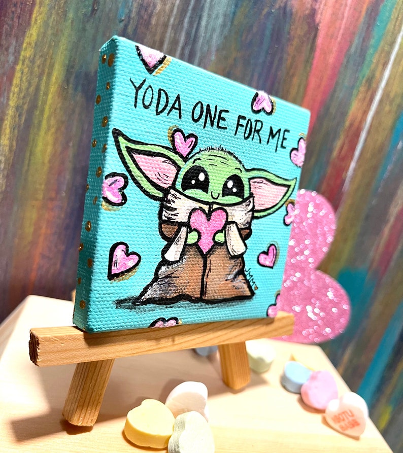 May include: A small, square canvas painting on a wooden easel. The artwork features a green character holding a pink heart, surrounded by pink hearts, with the text "YODA ONE FOR ME" on a light blue background.