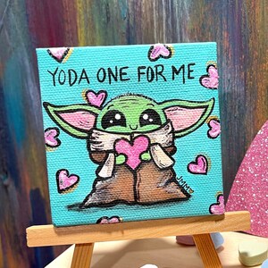 May include: A small square canvas painting featuring a cartoon depiction of Grogu, holding a pink heart. The painting has the text "YODA ONE FOR ME" and is surrounded by pink hearts on a teal background. The painting is displayed on a small wooden easel.
