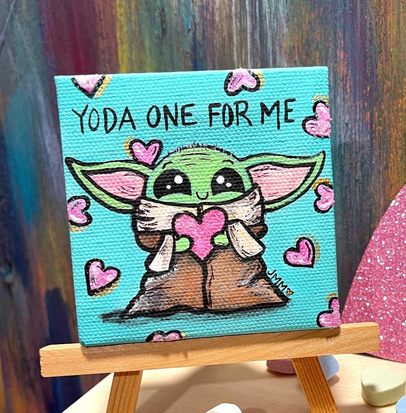 May include: A small square canvas painting featuring a cartoon depiction of Grogu, holding a pink heart. The painting has the text "YODA ONE FOR ME" and is surrounded by pink hearts on a teal background. The painting is displayed on a small wooden easel.
