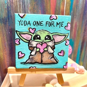 May include: A small, square canvas painting on a wooden easel. The artwork features a cartoon depiction of Grogu, holding a pink heart, with the text "YODA ONE FOR ME" on a light blue background. Pink hearts surround the character.