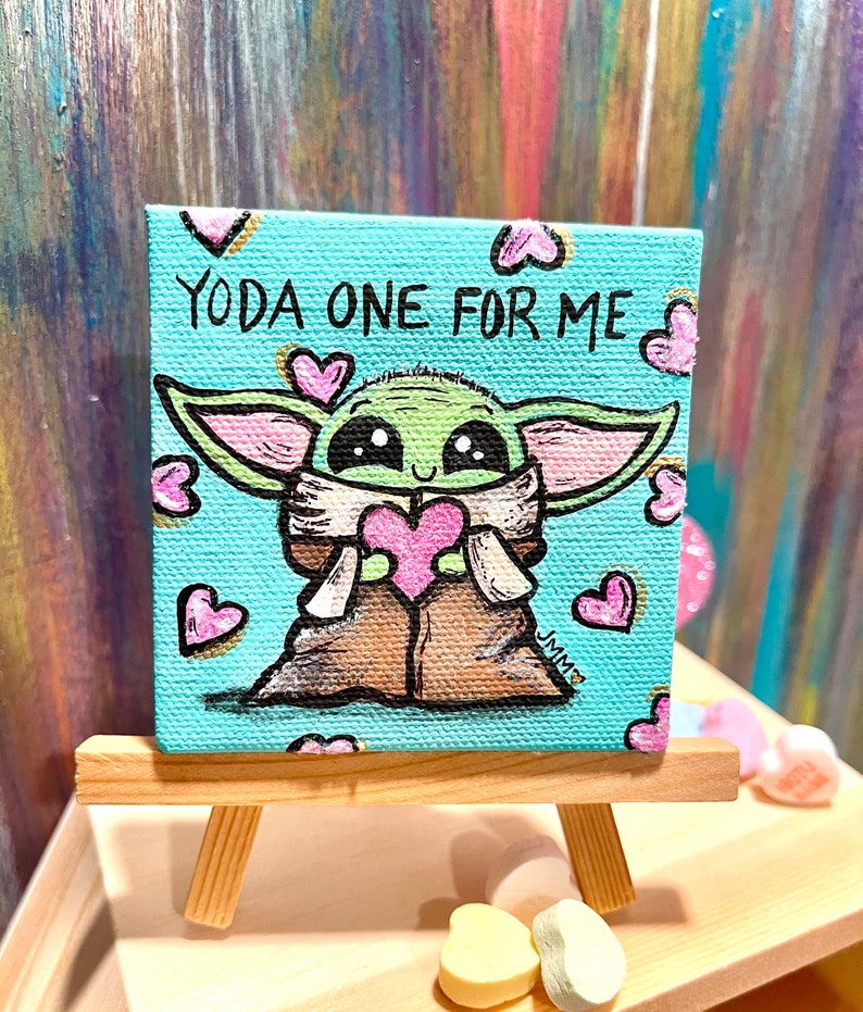 May include: A small, square canvas painting on a wooden easel. The artwork features a cartoon depiction of Grogu, holding a pink heart, with the text "YODA ONE FOR ME" on a light blue background. Pink hearts surround the character.