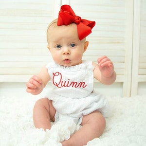 May include: A baby girl wearing a white dress with the name "Quinn" embroidered in red. She is wearing a red bow in her hair and is sitting on a white fluffy surface.