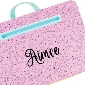 May include: Pink laptop stand with a blue handle and a built-in phone holder. The stand is decorated with a pattern of black and white corgi dogs and the name "Rimee" is written in black cursive.