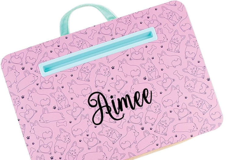 May include: Pink laptop stand with a blue handle and a built-in phone holder. The stand is decorated with a pattern of black and white corgi dogs and the name "Rimee" is written in black cursive.