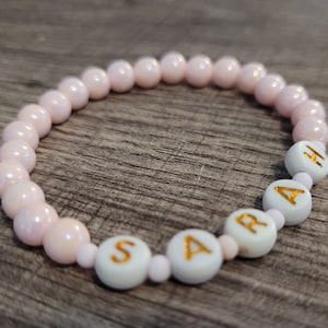 May include: A pink beaded bracelet with white beads that spell out the name "SARAH".