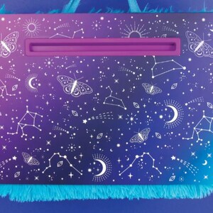 May include: A purple and blue laptop stand with a white fuzzy trim. The stand has a white design of stars, constellations, and butterflies.