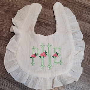 May include: White baby bib with a ruffled edge and green embroidered letters "PHE" with pink flowers.