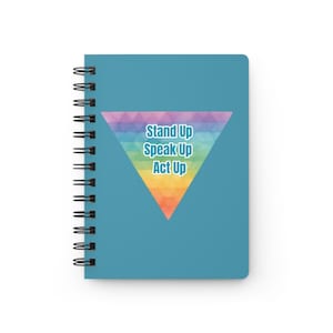 May include: A blue spiral-bound notebook featuring a large, downward-pointing triangle graphic composed of a multicolored geometric pattern. The text "Stand Up Speak Up Act Up" is printed in white letters with a blue outline in the center.